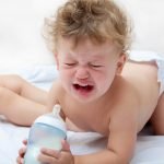 a small curly boy is standing on all fours, holding a bottle of milk and crying heavily