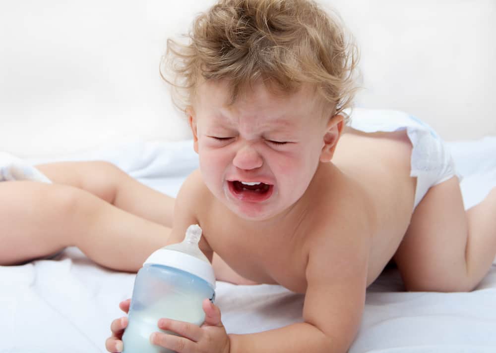 a small curly boy is standing on all fours, holding a bottle of milk and crying heavily