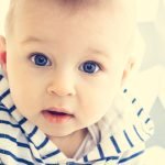 A cute white baby boy with big blue eyes is looking surprisingly into the camera and wearing striped hooded sweater.