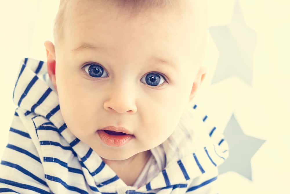 A cute white baby boy with big blue eyes is looking surprisingly into the camera and wearing striped hooded sweater.