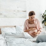 pregnant woman sits on bed and takes notes on laptop