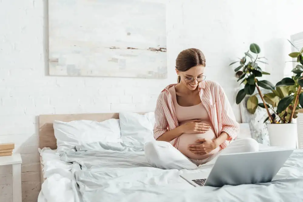 pregnant woman sits on bed and takes notes on laptop